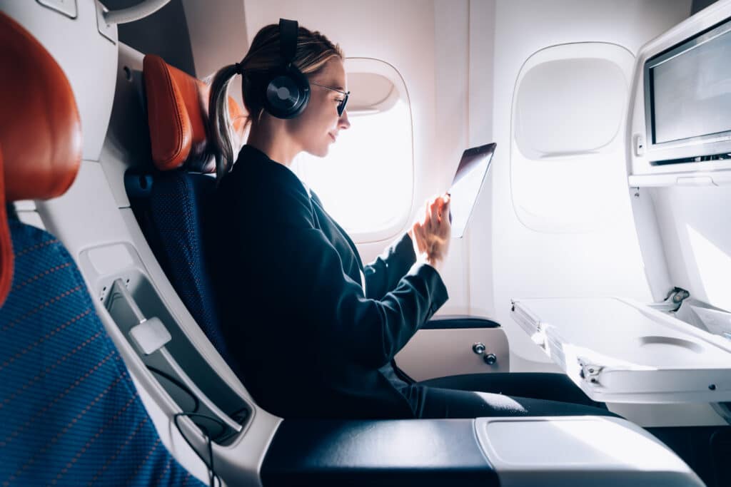 Eine entspannte Frau mit Kopfhörer und Tablet sitzt im Flugzeug.